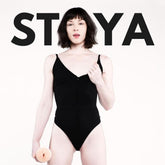A pale-skinned, dark-haired person in a black bodysuit stands holding an object, with "STOYA" in bold behind them, inspired by Fleshlight Girls: Stoya Destroya, available at Condomania.com.
